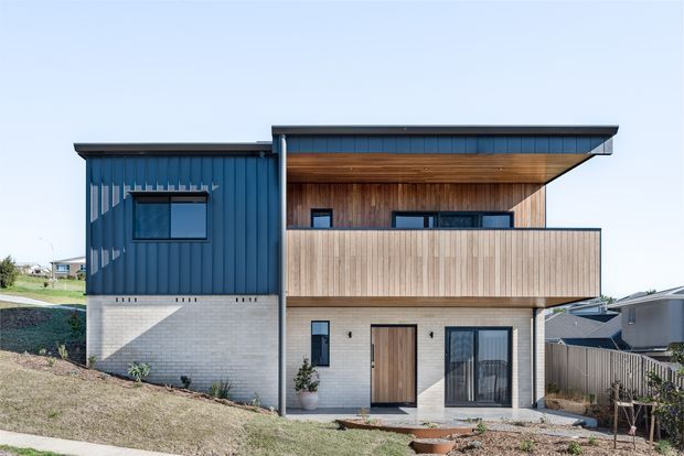 Exterior view of a contemporary two-storey residence on a sloping site. The facade combines dark blue vertical cladding, pale masonry at the lower level, warm timber-lined soffits and balcony privacy screening, with broad overhanging flat roof planes and modern glazing.