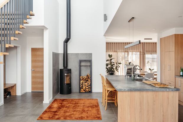 Bright open-plan contemporary interior combining a kitchen, dining area and entry/stair zone. A large timber island with stone benchtop anchors the kitchen, while a freestanding black wood-burning stove sits against a tiled feature wall beside stacked firewood. Vertical timber screening, timber cabinetry, a grey tiled floor, sculptural pendant lighting and a floating timber stair create a warm, refined material palette.