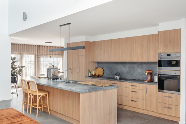 Bright contemporary open-plan kitchen with extensive light timber cabinetry, a large island with integrated sink and two bar stools, dark grey stone benchtops and splashback, and built-in stainless-steel ovens. Sheer curtains and dining furniture are visible in the adjoining area.
