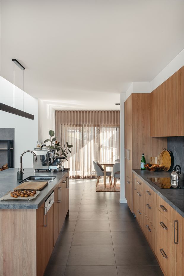 A bright, contemporary galley kitchen with extensive warm timber cabinetry, dark stone benchtops, integrated appliances and a central island with sink. A dining area is visible beyond the kitchen, framed by full-height sheer curtains and blinds. The space has large-format grey floor tiles, white walls and ceiling, and minimalist linear pendant lighting.