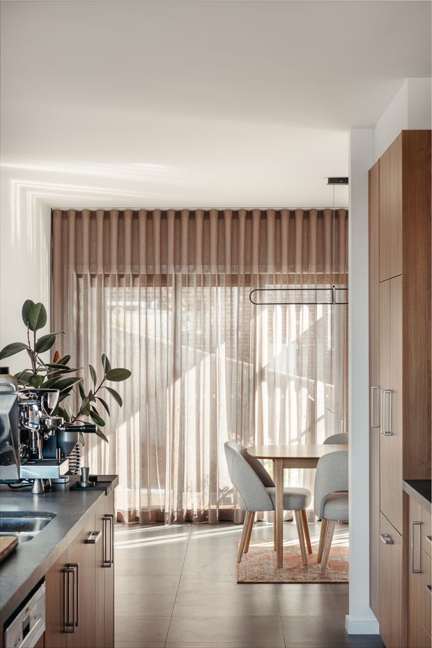 A bright, refined kitchen and dining area with warm timber cabinetry, a dark stone benchtop, stainless-steel appliances, and a compact dining table beside full-height sheer curtains. Soft daylight filters through the curtains, while indoor plants and contemporary pendant lighting add warmth and texture.