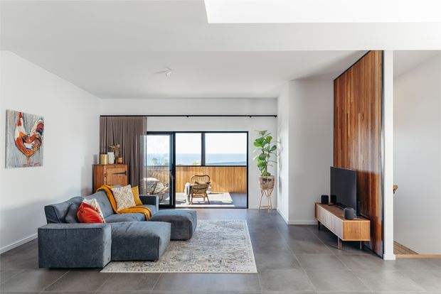 A bright contemporary living room with a large sectional sofa, patterned area rug, timber media wall and low console, and full-height sliding glass doors opening to a balcony. The room has white walls, dark grey tiled flooring, warm timber joinery, and views beyond the balcony.