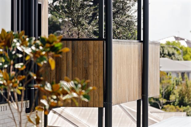 Close-up exterior view of a contemporary building featuring dark slender metal posts, large glazed openings, and warm vertical timber wall cladding. Landscaped greenery and neighbouring rooflines are visible in the background.