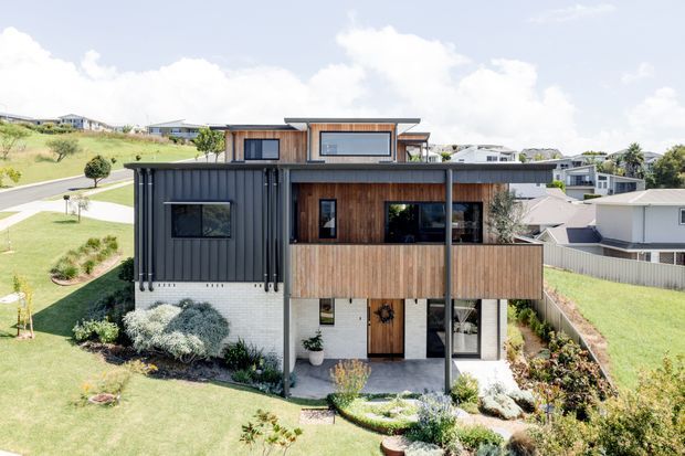 Modern multi-level residential house on a sloping suburban site, viewed from the landscaped front garden. The facade combines dark vertical cladding, warm timber screening and white masonry, with broad glazing, projecting balconies and a low-pitched flat roof form.