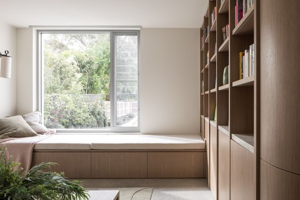 A calm, light-filled built-in window-seat nook with upholstered cushioning and extensive floor-to-ceiling timber shelving and cabinetry. The large window looks onto leafy greenery, while neutral walls, warm wood joinery, and soft textiles create a refined reading or living area.