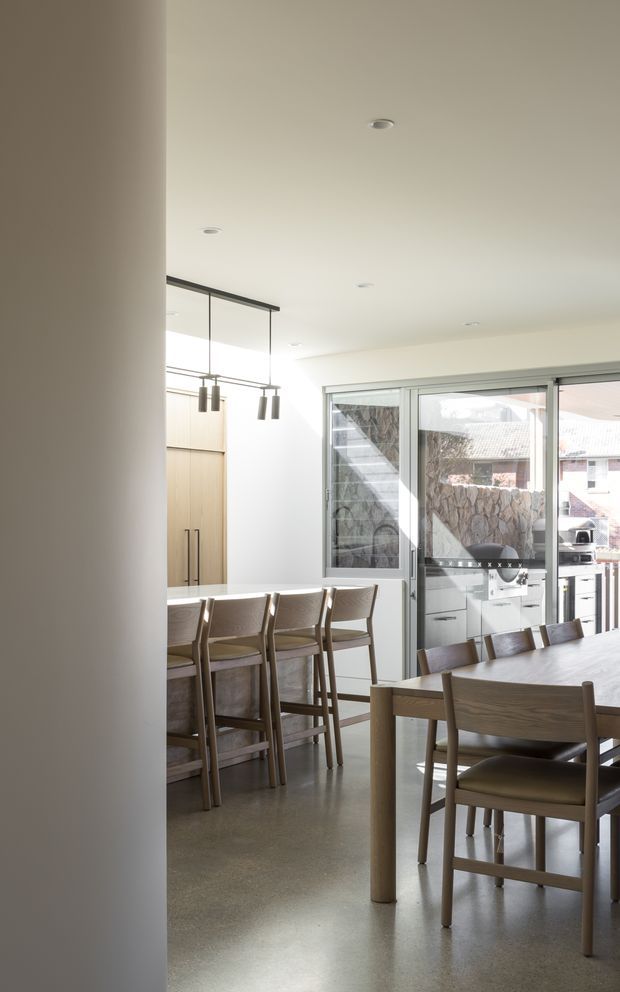 A bright, contemporary open-plan dining and kitchen area with a long timber dining table and matching chairs in the foreground, a kitchen island with timber cabinetry and bar stools to the left, and large sliding glass doors opening to an outdoor entertaining area. The space has a smooth light ceiling with recessed downlights, a polished concrete floor, minimalist linear pendant lighting, and neutral warm-toned finishes.