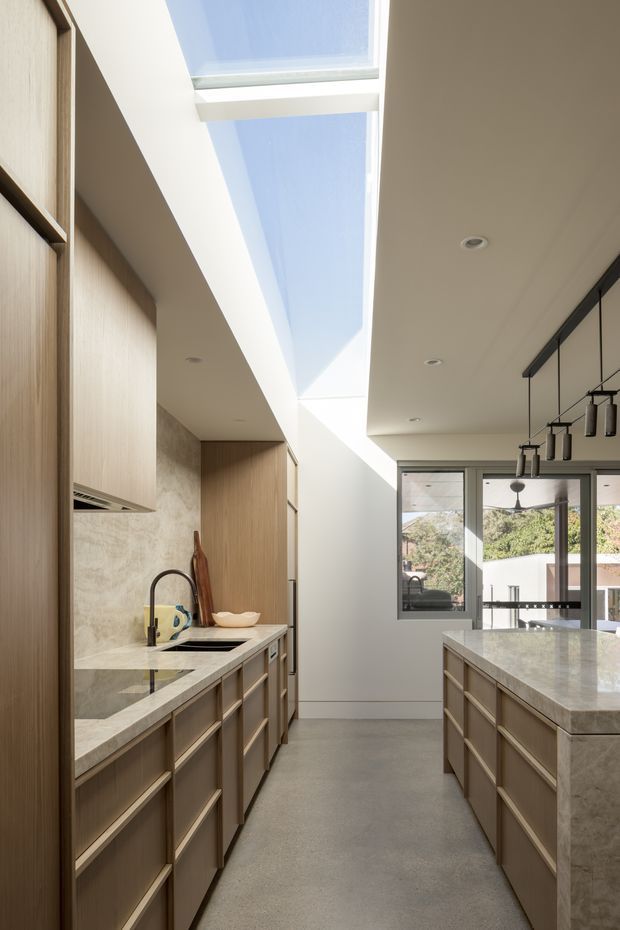 A bright, contemporary galley kitchen with a central island and extensive light timber cabinetry. The kitchen features pale stone benchtops and matching stone wall cladding, integrated appliances, a black gooseneck tap, and a row of black pendant lights. A dramatic linear skylight brings daylight into the space, while glazing at the rear provides views to greenery outside.