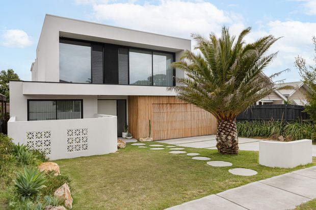 Exterior view of a contemporary two-storey residence with a crisp white rendered facade, strong rectilinear forms, dark-framed glazing, a timber-screened garage or entry facade, and a landscaped front garden with stepping stones and a palm tree.