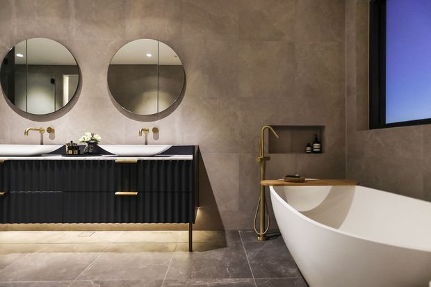 Luxurious contemporary bathroom featuring a freestanding sculptural bathtub, large wall-mounted black fluted vanity with brass hardware, twin round mirrors, wall-mounted gold-toned fixtures, recessed shelving, and large-format stone-look wall and floor finishes. A dark-framed window adds contrast.