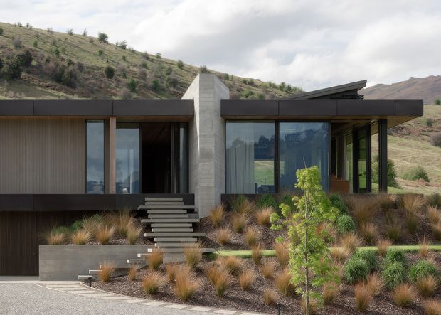 A low, modern single-storey house set into a dry, sloping hillside landscape. The facade combines dark horizontal cladding, large glazed openings, a prominent smooth concrete central volume, and a sharply angled roof plane. Floating steps lead through a landscaped front garden with ornamental grasses and native-style planting.