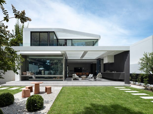 Contemporary two-storey residence viewed from the landscaped front garden, featuring a crisp white facade, extensive glazing, a curved corrugated metal roofline, deep cantilevered outdoor living terrace, and an integrated dark outdoor kitchen or bar area. The garden includes clipped shrubs, gravel beds, lawn, and stepping stones.