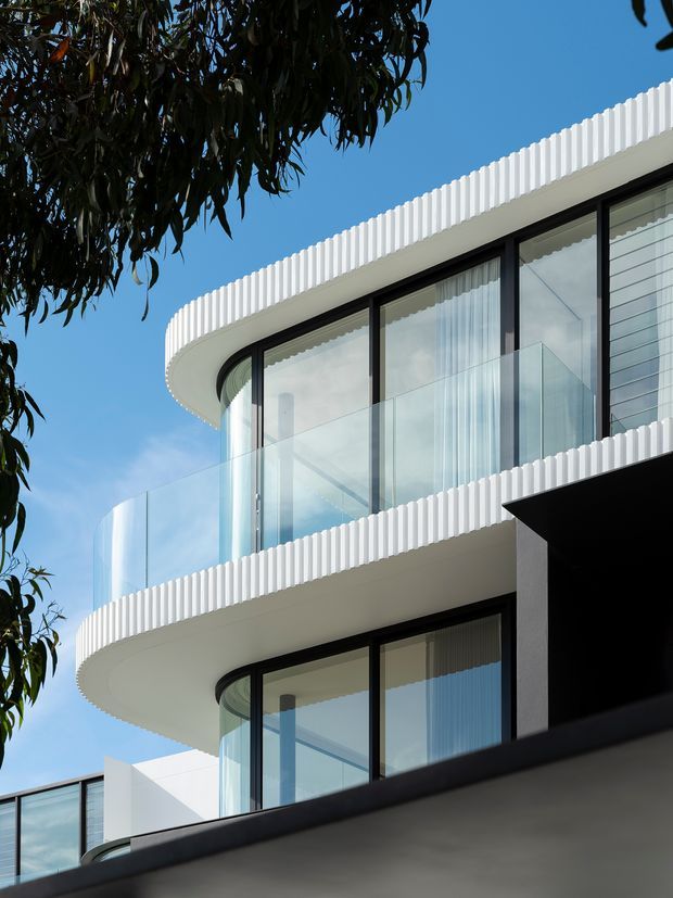 Upward-looking exterior view of a contemporary multi-storey building with rounded projecting balconies, extensive floor-to-ceiling glazing, transparent glass balustrades, and distinctive white vertically ribbed facade and balcony edges. Dark window frames contrast with the bright white exterior, with tree foliage and blue sky framing the building.