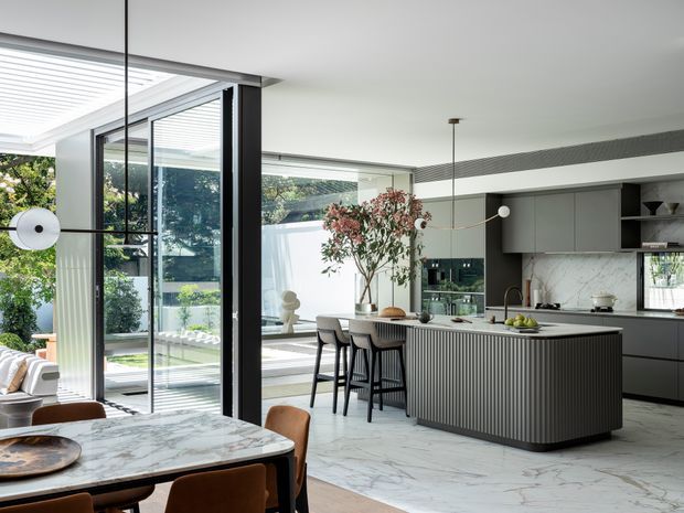Bright, open-plan contemporary kitchen and dining area with a large fluted island, dark cabinetry, pale marble-look flooring and benchtops, floor-to-ceiling black-framed glazing, and an adjoining living space. The room opens toward a landscaped outdoor courtyard, with sculptural pendant lighting and a dining table in the foreground.