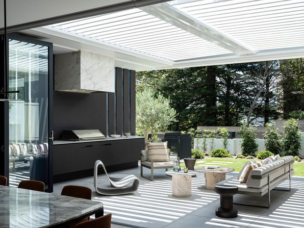 A bright contemporary indoor-outdoor living and dining space opening onto a landscaped patio. The room features a large marble dining table, dark built-in cabinetry with marble backsplash, sculptural lounge seating, a sectional sofa, and a white slatted pergola or glazed roof creating strong linear shadows. Floor-to-ceiling glazing connects the interior to the garden.