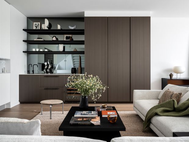 A refined open-plan living room with a contemporary dark timber built-in joinery wall, integrated kitchenette or bar area, open display shelving, and a large upholstered sectional sofa. A black rectangular coffee table, textured rug, sculptural decor, and soft neutral furnishings create a sophisticated, calm interior. A mirrored backsplash reflects the room and shelving, while full-height cabinetry provides concealed storage.