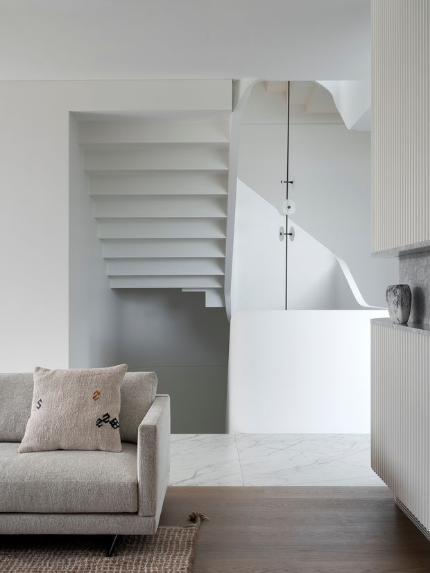 A refined, monochromatic interior featuring a pale upholstered sofa in the foreground, a white sculptural staircase with open shelving, a concealed white door, and a curved white partition. The space combines white marble-look flooring with warm timber flooring, fluted built-in cabinetry, and subtle decorative objects. The overall effect is minimalist, calm, and highly sculptural.
