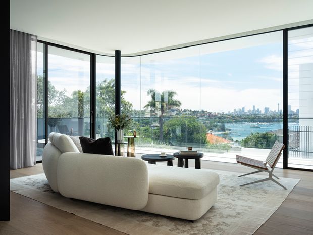 Bright contemporary living room with a sculptural cream sectional sofa, round coffee tables, timber flooring, and full-height curved glazing overlooking a harbour, waterfront homes, and a distant city skyline. A black structural column, sheer curtains, and a timber-framed lounge chair complete the refined interior.
