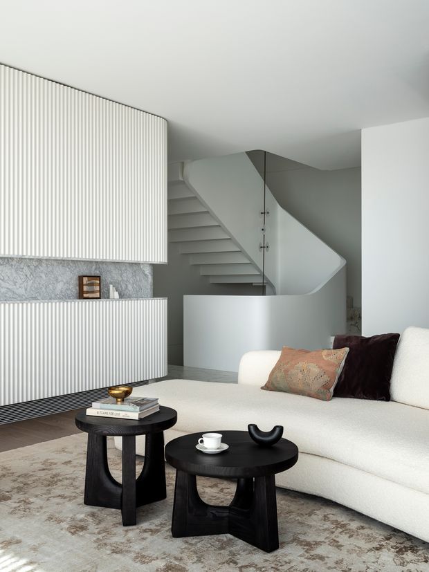 Refined contemporary living room with a large cream boucle sofa, black sculptural nesting coffee tables, patterned rug, and neutral decorative cushions. The room features a dramatic sculptural staircase with a curved balustrade and floating steps, plus ribbed white built-in cabinetry and a stone-textured wall.