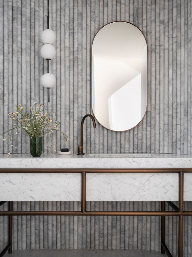 A refined bathroom vanity vignette featuring a fluted grey wall, marble countertop and drawer fronts, a slim bronze-toned metal frame, an arched oval mirror, and sculptural globe wall lighting. A dark bronze curved faucet and small decorative vase complete the calm, luxurious composition.