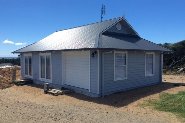 Single-storey light-blue weatherboard building with a broad corrugated metal gable roof, white-framed windows, a small decorative gable vent, and exterior wall lantern. The building sits on a mostly bare sandy site with limited surrounding vegetation.