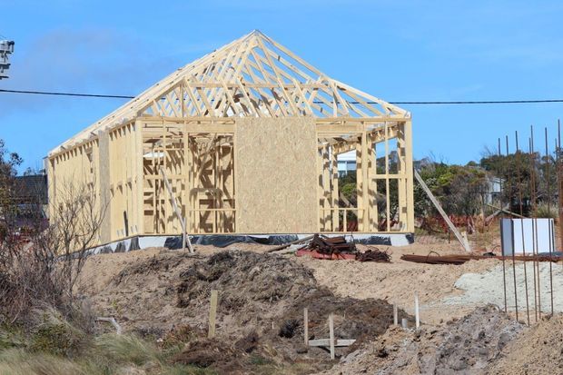 Exterior construction photograph showing a timber-framed house under construction on a dirt building site. The pitched gable roof structure, exposed light timber studs and rafters, partial sheet bracing, concrete foundation, and surrounding construction materials are clearly visible under a blue sky.
