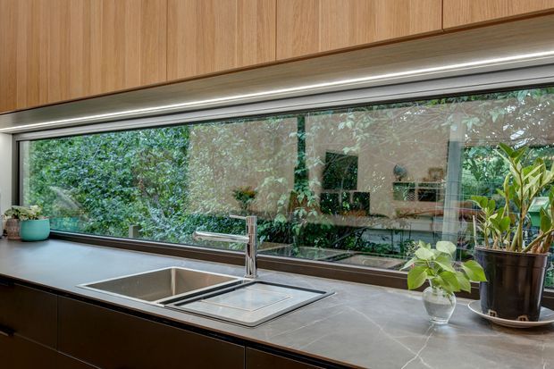 Contemporary kitchen work area featuring a long dark stone benchtop with an integrated stainless-steel sink and drainer, timber cabinetry above and below, and a large horizontal glazed window overlooking lush greenery. Under-cabinet lighting and indoor plants add warmth and texture.