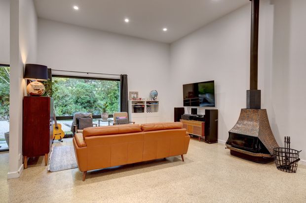 A bright, high-ceilinged living room with white walls and recessed downlights. The room features a large orange-brown leather sofa, a timber media console and television, a tall timber cabinet, shelving, and a freestanding sculptural fireplace with a dark metal flue and textured hood. Large glazing provides views of dense greenery outside, while a rug, armchair, and small decorative furnishings complete the space.