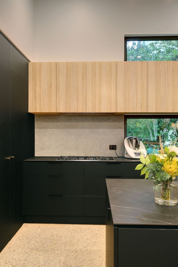 A compact contemporary kitchen with matte black lower cabinetry and tall pantry joinery, natural light-oak overhead cabinets, a warm grey stone-look splashback, dark benchtops, and a terrazzo-style floor. Large windows overlook leafy greenery, while a peninsula provides additional work surface. A freestanding countertop appliance and flower arrangement are visible.