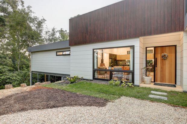 A contemporary single-storey house exterior set among mature trees, with a contrasting dark timber-clad upper facade, pale horizontal weatherboard-style cladding, a broad glazed living area, and a timber entry door. A gravel forecourt and stepping-stone path lead to the entrance, while the interior kitchen and living space are visible through the window.