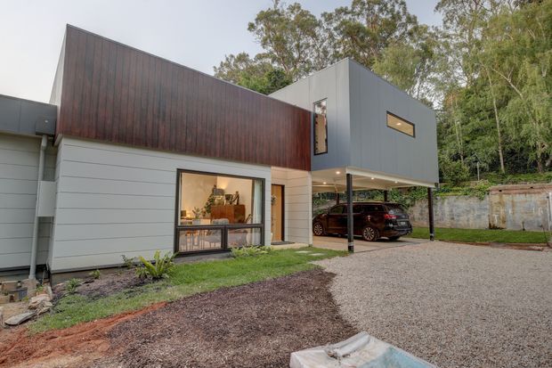 Exterior view of a contemporary two-storey residence with a sculptural gabled roof form, dark vertical timber cladding, light grey horizontal wall panels, and a projecting upper level over a covered carport. Large windows reveal a warmly furnished interior, while gravel paving, landscaping, mature trees, and a retaining wall surround the home.
