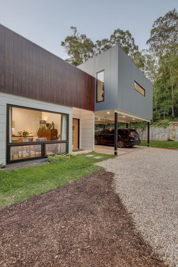Exterior view of a contemporary two-storey house with a cantilevered upper volume, dark vertical timber cladding, grey metal-like wall panels, extensive glazing, and a covered carport. The home sits beside a gravel driveway and landscaped garden, with mature trees and a masonry boundary wall behind it.