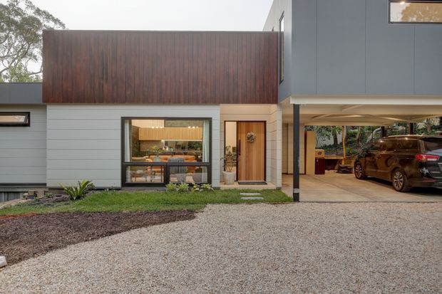 Contemporary residential exterior with a low, horizontally composed form, dark vertical timber cladding above pale horizontal wall cladding, and a warm timber entry door. Large glazing reveals a furnished interior, while a covered carport occupies the right side. The gravel forecourt and restrained landscaping reinforce the modern, natural-material aesthetic.