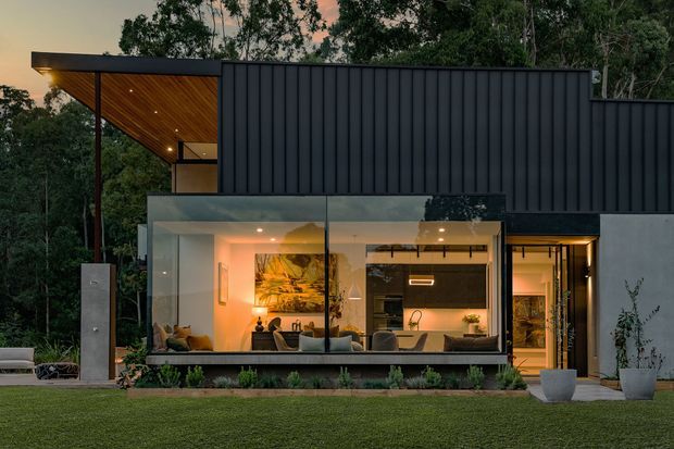 Contemporary single-storey residential exterior at dusk, viewed from a landscaped lawn. The home combines dark vertical metal cladding with broad floor-to-ceiling glazing, revealing a warmly lit open-plan living area inside. A projecting timber-lined roof canopy with recessed lighting creates a sheltered outdoor threshold, while the clean rectilinear form and integrated landscaping give the house a refined modern character.