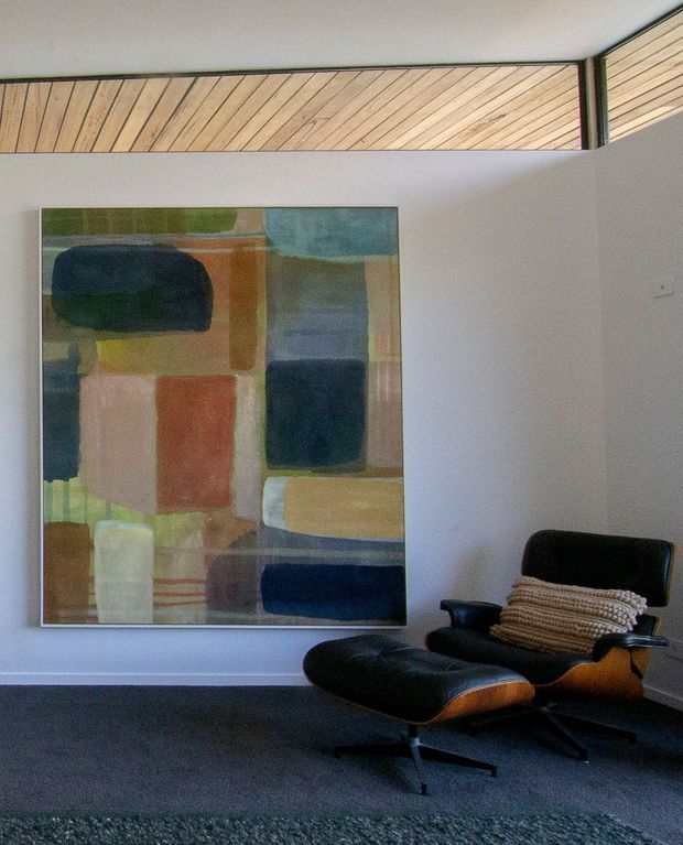 A quiet, mid-century-inspired sitting area with a black leather lounge chair and matching ottoman positioned against a pale wall. A large abstract geometric artwork adds muted blue, green, ochre, terracotta, and cream tones. The room features dark carpeting and a distinctive timber-lined clerestory or angled ceiling recess with slim dark framing.