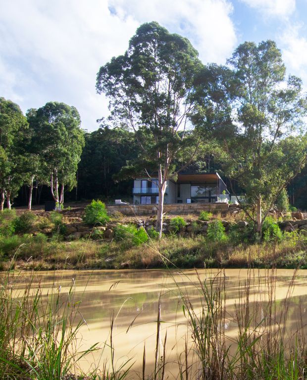 A small contemporary house set among dense mature trees on a gently elevated, rocky site. The low rectangular dwelling has dark cladding, extensive glazing, and a simple pitched roof, with a stone retaining wall and landscaped garden in front. A calm pond or waterway with reeds occupies the foreground.