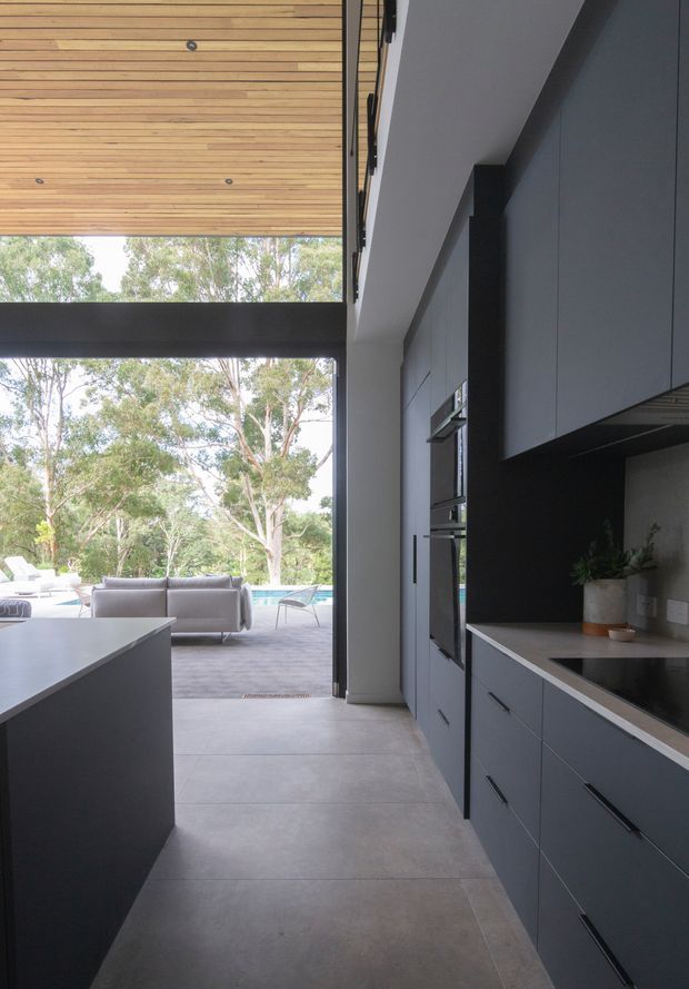 A contemporary kitchen viewed along a narrow circulation zone toward a glazed outdoor entertaining area. The kitchen features dark matte built-in cabinetry with integrated appliances, a pale stone benchtop, large-format grey floor tiles, and a substantial dark island. A timber-lined ceiling and black structural framing surround full-height glazing, with a pool and leafy landscape visible beyond. The space is clean-lined, spacious, and strongly connected to the outdoors.