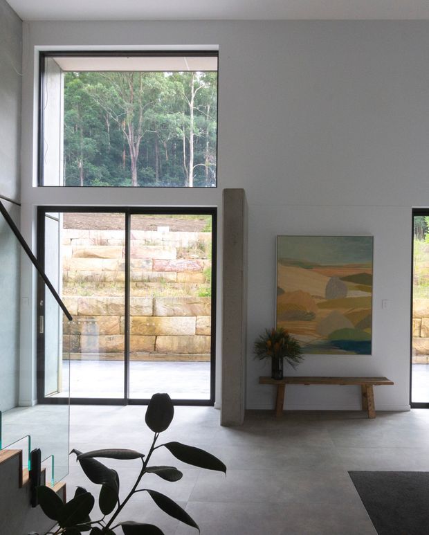 A bright, minimalist interior with a tall glazed opening and black-framed sliding doors overlooking a landscaped stone retaining wall and wooded setting. The space has smooth pale walls, a light grey tiled or concrete-look floor, a slim timber console bench, abstract landscape artwork, and sparse contemporary furnishings. A partial stair or dark structural element appears at the left edge, suggesting an open living or entry area.