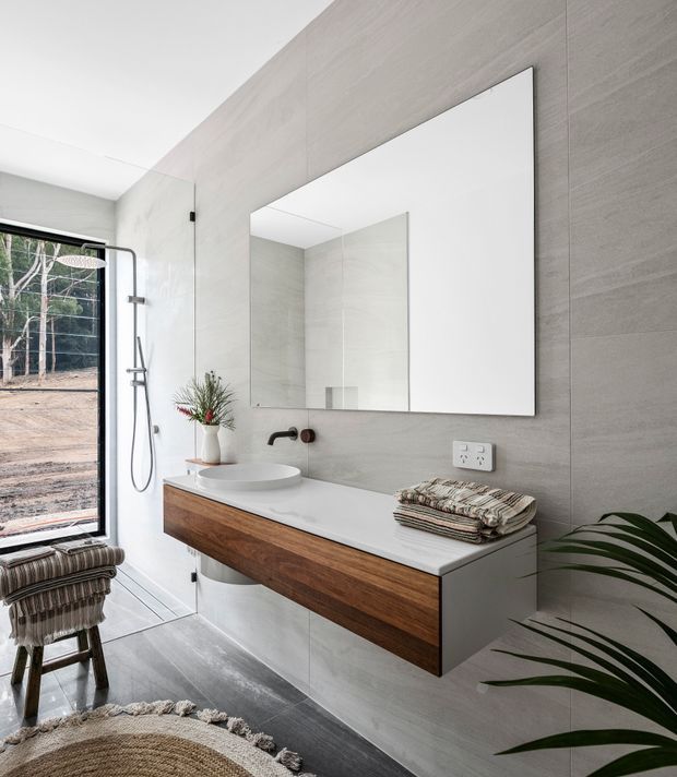 Bright contemporary bathroom with a floating timber vanity, white integrated benchtop and vessel basin, wall-mounted dark tapware, large frameless mirror, and grey full-height tiled walls. A frameless glass shower enclosure with exposed shower hardware sits beside a large window overlooking trees. Layered textiles, a woven rug, and indoor plants add warmth and an organic touch.
