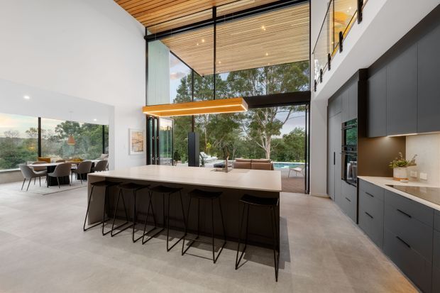 A spacious contemporary kitchen with a large island and four black bar stools, dark built-in cabinetry, integrated appliances, and a pale stone benchtop. The kitchen opens to a dining area and an outdoor terrace through extensive floor-to-ceiling glazing. A timber-lined ceiling, black-framed glazing, double-height volume, and a linear pendant light create a refined indoor-outdoor setting.