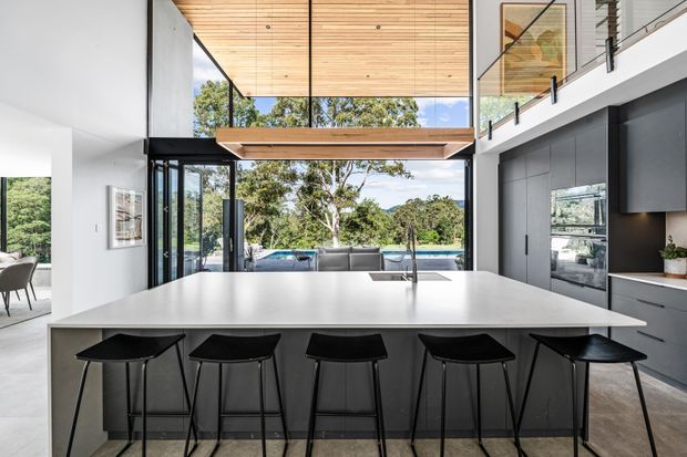 Bright contemporary open-plan kitchen dominated by a large white island with integrated sink and seating for five on black bar stools. Dark grey full-height cabinetry lines the right wall, while extensive floor-to-ceiling glazing opens the space toward a pool and landscaped garden. A timber-lined ceiling, double-height volume, black-framed glazing and a glass-balustraded upper-level walkway create a refined indoor-outdoor composition.