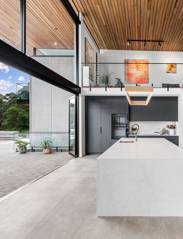 A dramatic open-plan contemporary kitchen in a double-height space, featuring a large white island, dark matte cabinetry, integrated appliances, expansive black-framed glazing, a timber slatted ceiling, and a mezzanine-style upper level with glass balustrades. The kitchen opens directly to a paved outdoor terrace with greenery visible beyond.