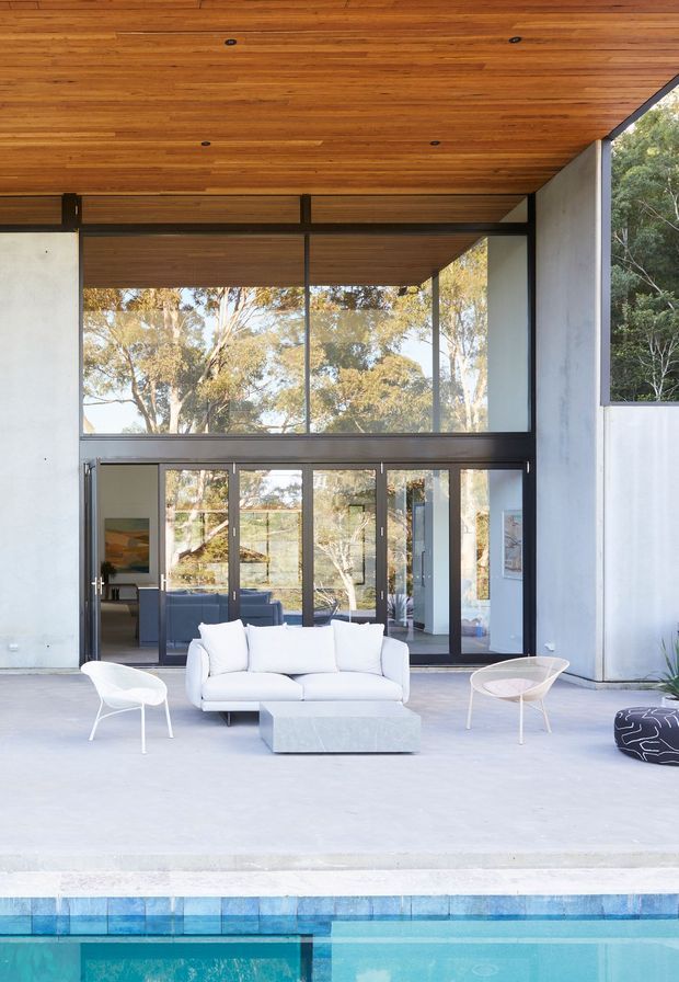 A bright, modern indoor-outdoor living space opening directly onto a swimming pool. The room features a white sofa, sculptural lounge chairs, a low rectangular coffee table, expansive dark-framed glazing and glass doors, pale concrete flooring, and a warm timber-lined ceiling. Mature trees are visible through the glazing, creating a strong connection to the landscaped setting.