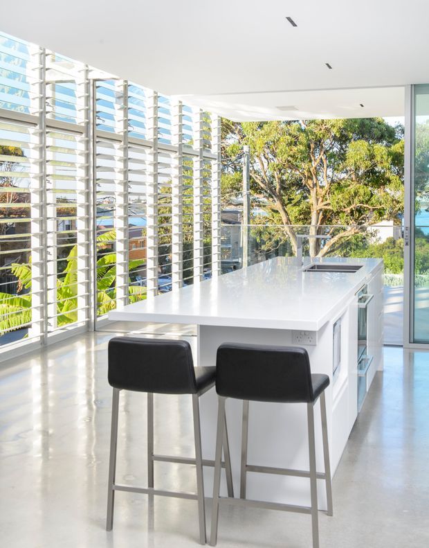 Bright contemporary kitchen interior with a large white island and integrated sink, two black bar stools, polished light flooring, extensive floor-to-ceiling glazing, and horizontal external louvre screens. Mature trees and neighbouring buildings are visible outside.