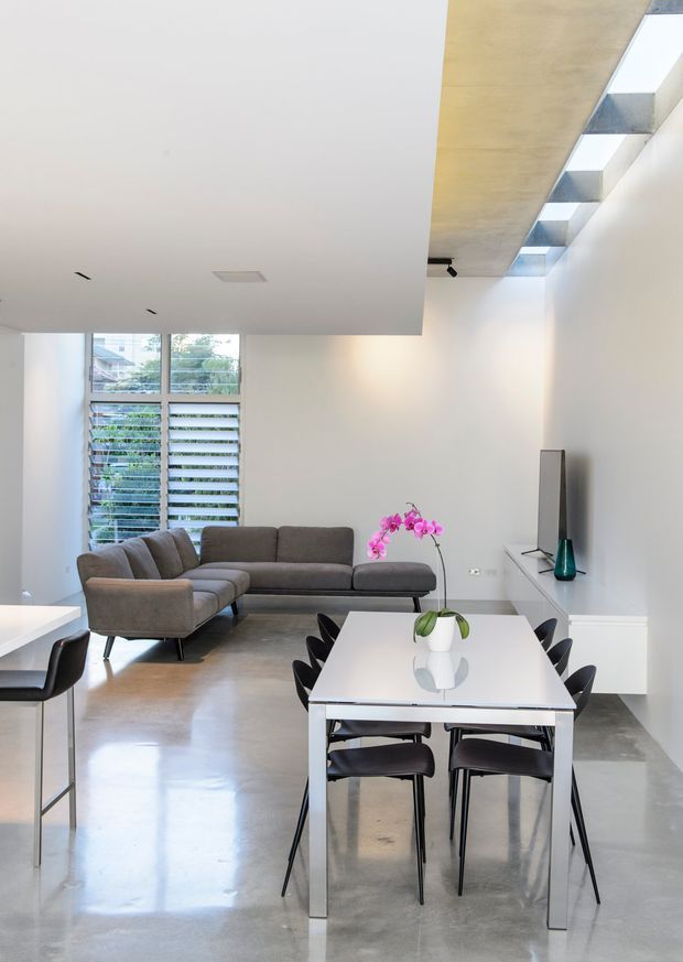 Bright open-plan contemporary living and dining area with a polished concrete floor, white walls and ceilings, a grey sectional sofa, white dining table with black chairs, and minimalist built-in media cabinetry. A large louvred window brings in natural light, while a ceiling-level skylight or clerestory adds additional illumination.