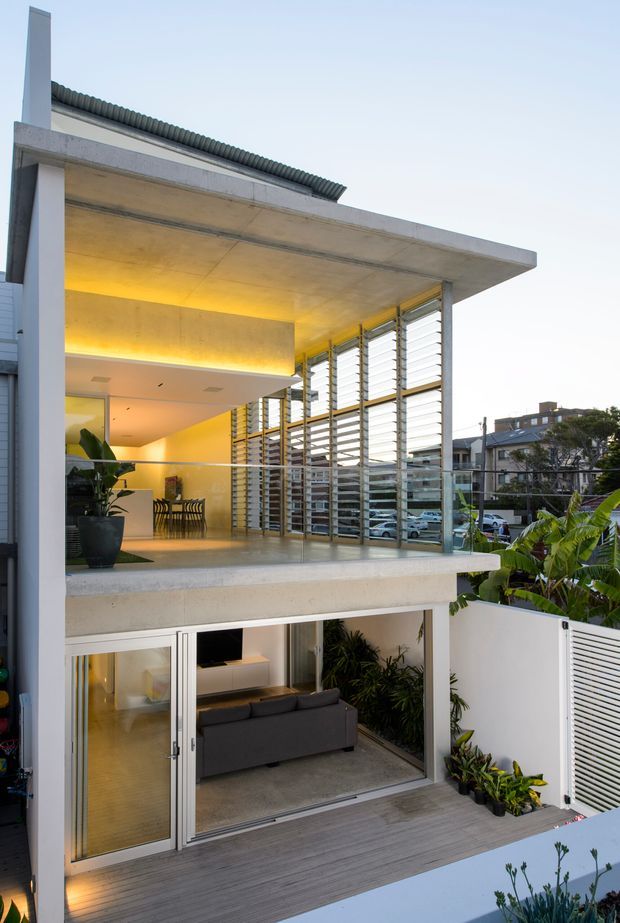 Exterior view of a contemporary multi-level residence with exposed concrete slabs, broad cantilevered roof forms, extensive louvred glazing, and glass balustrades. A warm-lit upper-level living and dining area opens onto a balcony, while the lower level has full-height glazed doors opening to a small landscaped outdoor area. The facade combines concrete, metal louvres, glazing, and planted terraces.