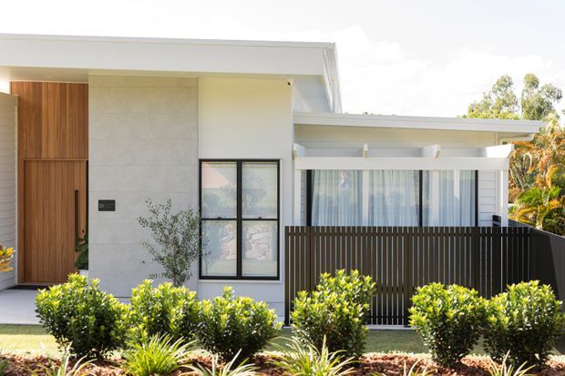 Contemporary single-storey residential facade with crisp white rendered walls, a large timber pivot entry door, pale stone or concrete wall cladding, black-framed glazing, and a covered front porch screened by a dark vertical slatted fence. Neatly clipped shrubs and ornamental planting form the foreground landscape.