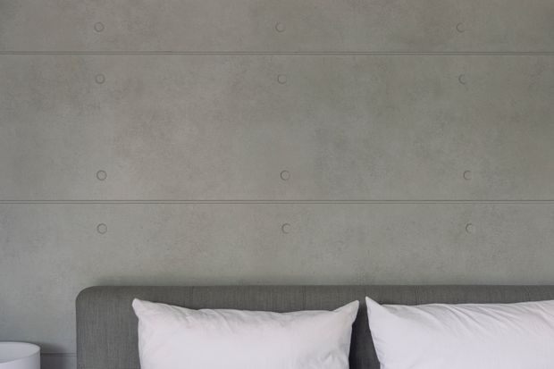 Close-up of a minimalist bedroom headboard with two white pillows against a grey upholstered bed frame. The background wall is finished in smooth grey concrete with visible horizontal panel joints and circular form-tie marks, creating an understated industrial look.