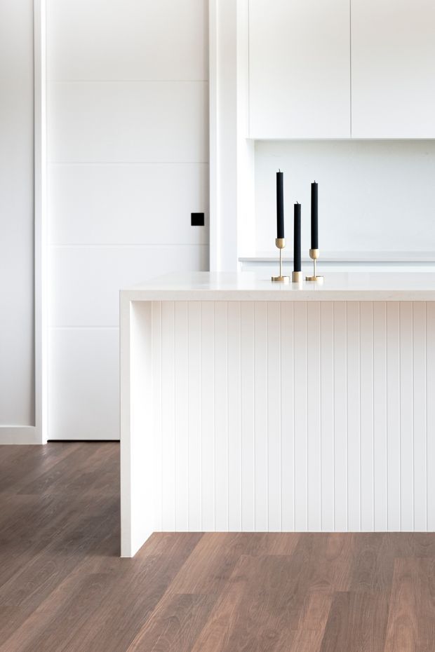 Bright minimalist kitchen interior featuring a white island with vertical wall panelling, integrated white cabinetry, a flush-panel door, dark timber flooring, and three black taper candles with brass holders on the benchtop. The composition is clean, restrained, and high-contrast with strong white surfaces against warm wood flooring.