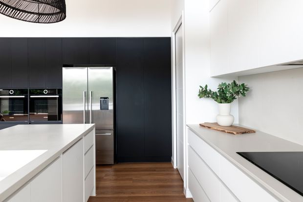 Contemporary kitchen with a large white island, extensive white cabinetry, matte black full-height cabinetry, integrated ovens, stainless-steel double-door refrigerator, dark timber flooring, and a black induction cooktop. A woven pendant light and simple plant styling add warmth and texture.