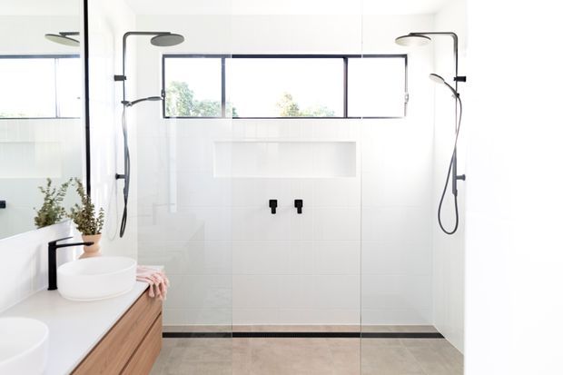 Bright contemporary bathroom with a frameless glass walk-in shower, dual black shower fixtures, white tiled walls, clerestory-style windows, a recessed wall niche, and a timber vanity with white vessel basins. The space has a clean, minimal palette with black accents and natural wood joinery.